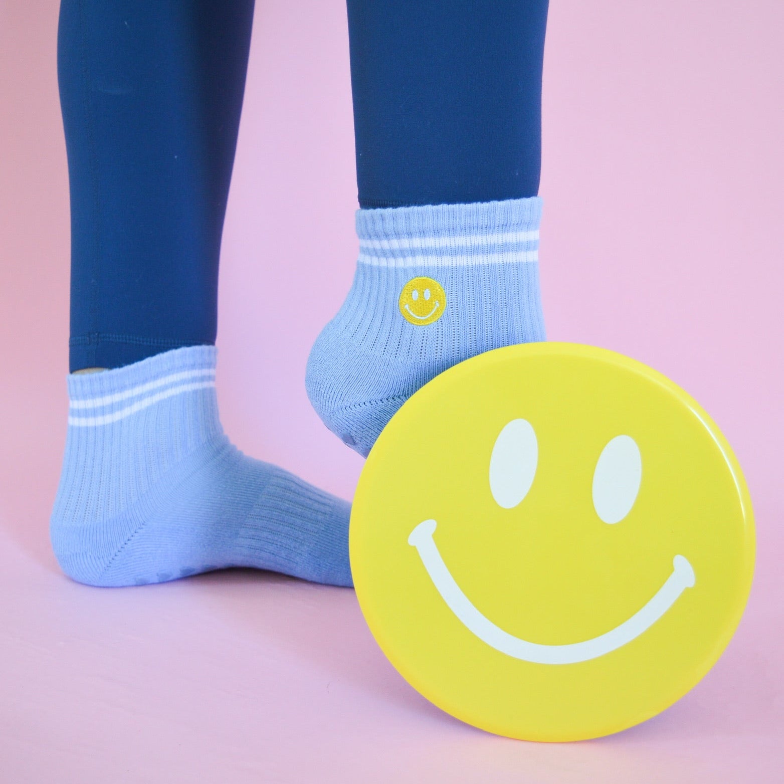 Person wearing light blue socks with a smiley face design, standing next to a yellow smiley face sliding disk on a pink background.