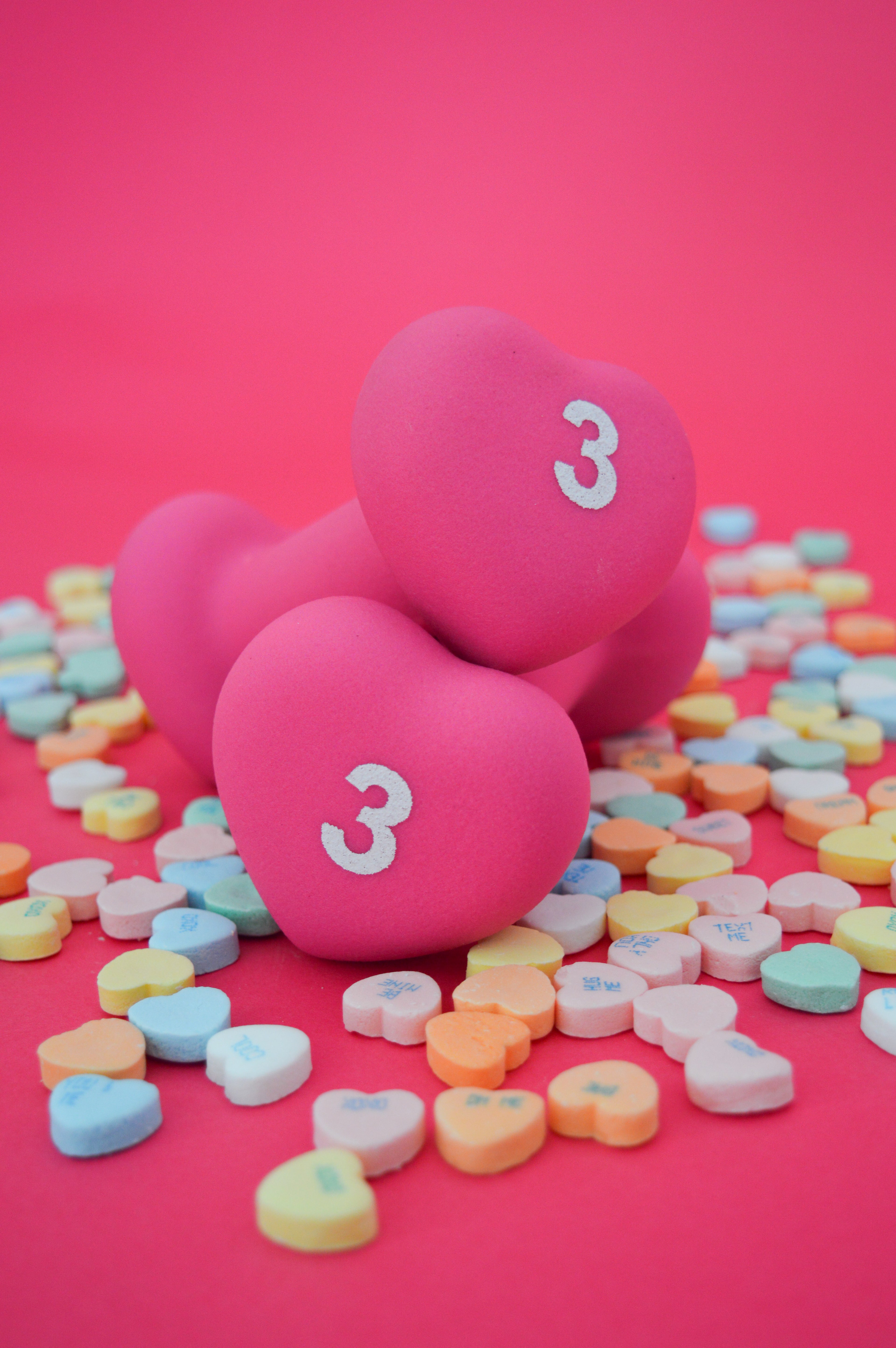 Pink heart-shaped dumbbells with the number 3 on a pink background with colorful heart-shaped candies.