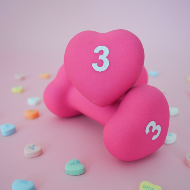 Pink dumbbells 3lb on a pink background with colorful heart-shaped candies.