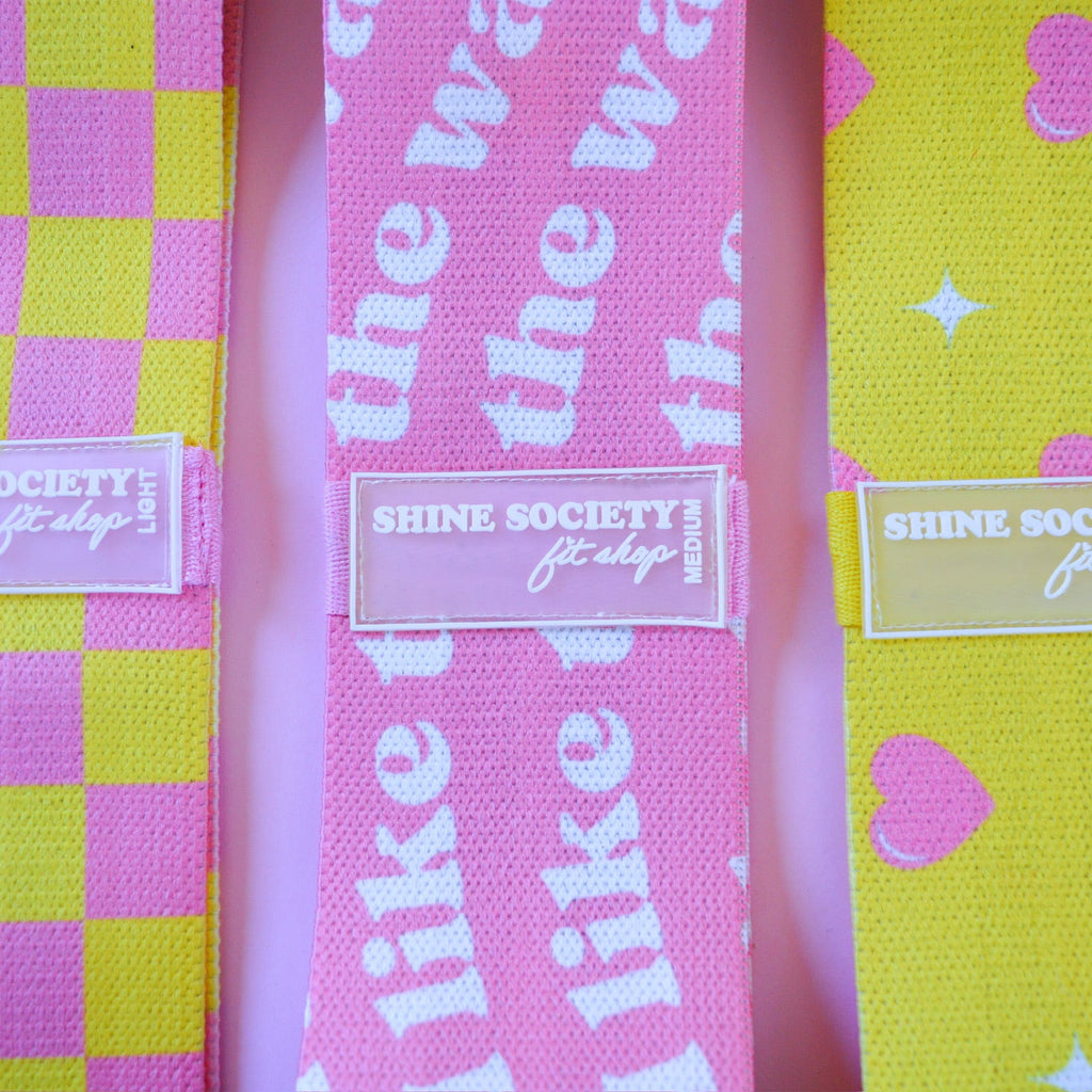 Three colorful Hip bands with different patterns and 'Shine Society' labels on a pink background.